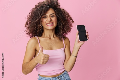 Woman blogger holding phone video call takes selfies, with curly hair in pink smile t-shirt and jeans poses on pink background, copy space, technology and social media, online