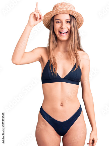 Young beautiful girl wearing bikini and hat pointing finger up with successful idea. exited and happy. number one.