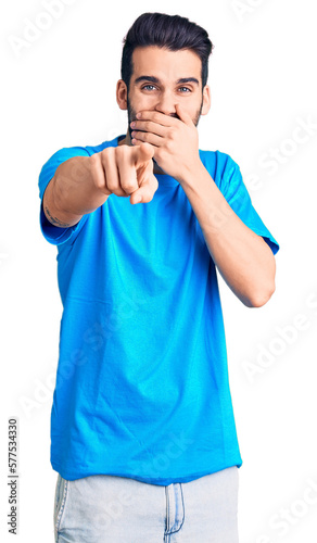 Young handsome man with beard wearing casual t-shirt laughing at you, pointing finger to the camera with hand over mouth, shame expression