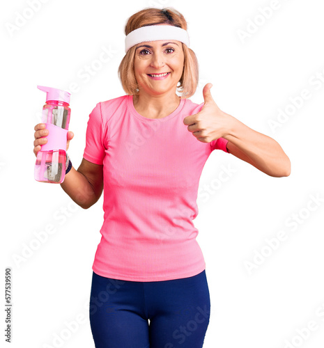 Young blonde woman wearing sportswear holding water bottle smiling happy and positive, thumb up doing excellent and approval sign © Krakenimages.com