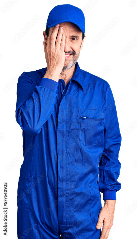 Middle age handsome man wearing mechanic uniform covering one eye with hand, confident smile on face and surprise emotion. Middle age handsome man wearing mechanic uniform covering one eye with hand, confident smile on face and surprise emotion.