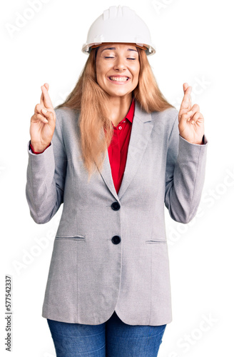 Beautiful young woman wearing architect hardhat gesturing finger crossed smiling with hope and eyes closed. luck and superstitious concept. © Krakenimages.com
