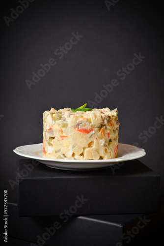 Side view of Traditional Russian salad Olivier served on white plate located on black box. Salad with potato and carrot cubes, green peas, pickled cucumbers, eggs with spring onions on Dark background