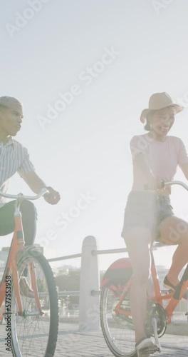 Vertical video of happy biracial couple hriding bikes on promenade, in slow motion photo