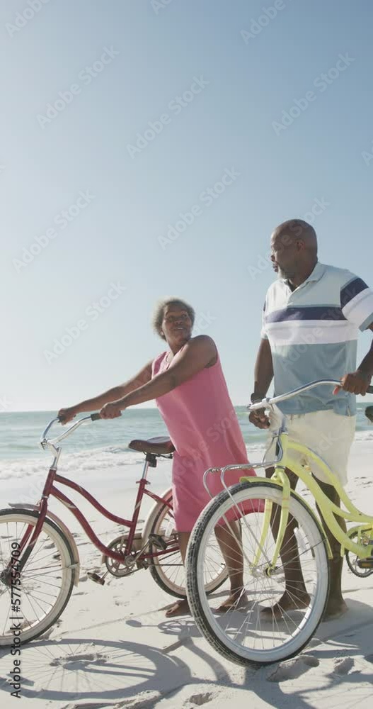 Vertical video of senior african american couple walking with bikes at beach, in slow motion