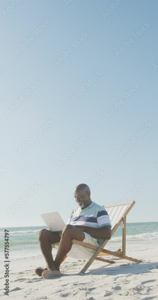 Vertical video of senior african american man using laptop on deckchair at beach, in slow motion Vertical video of senior african american man using laptop on deckchair at beach, in slow motion