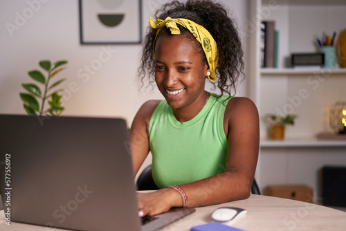 Young African smiling woman using laptop in home office. Happy girl typing on computer and telecommuting. Cheerful people sitting at desk in studio. Concept of enterprising and hardworking person. 