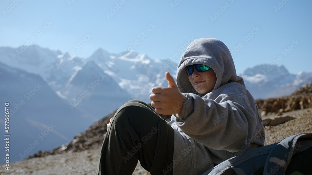 Boy gives thumbs up in mountains. Creative. Boy in ski goggles looks at mountain landscape on sunny day. Beautiful mountain landscape and enjoying boy