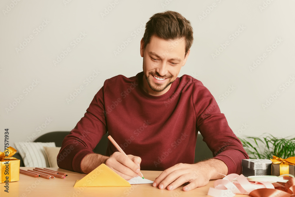 Happy man writing message in greeting card at wooden table in room Happy man writing message in greeting card at wooden table in room
