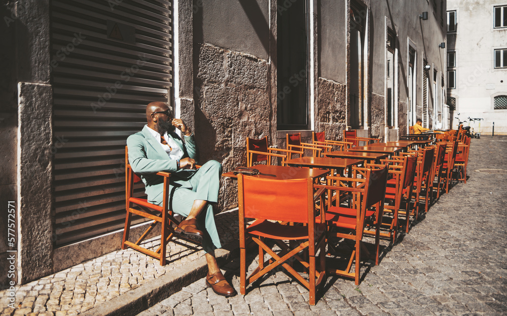 Naklejka premium A stylish Black man bald-headed sits alone, lost in thought, on a closed terrace in Lisbon, Portugal; the outdoor furniture remains, shining in the bright sunshine against the traditional pavement