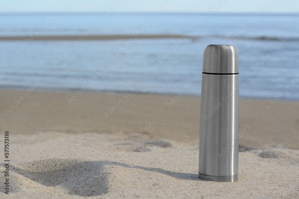 Metallic thermos with hot drink on sandy beach near sea, space for text Metallic thermos with hot drink on sandy beach near sea, space for text