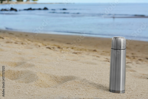 Metallic thermos with hot drink on sandy beach near sea, space for text