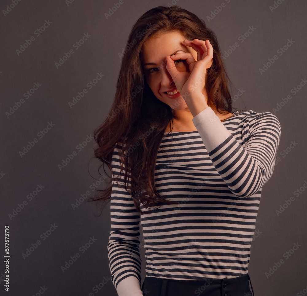 Happy excited business brunette woman doing spyglass sign looking in fingers near the eye in stripped casual t-shirt. Concept studio portrait. Vintage toned color Happy excited business brunette woman doing spyglass sign looking in fingers near the eye in stripped casual t-shirt. Concept studio portrait. Vintage toned color