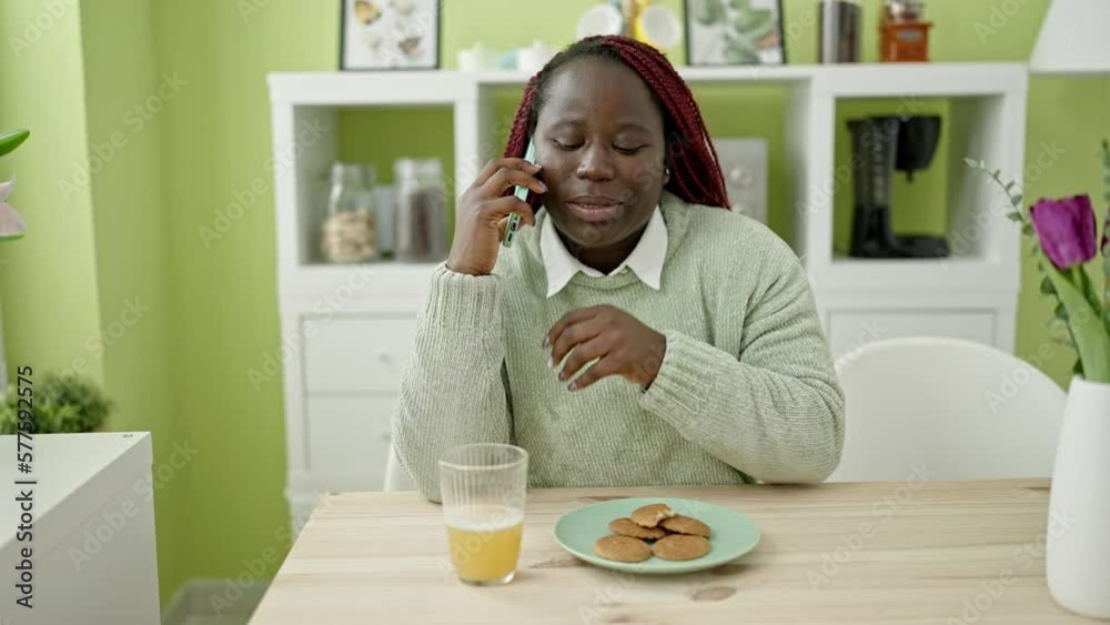 African woman with braided hair eating cookies using smartphone at dinning room African woman with braided hair eating cookies using smartphone at dinning room