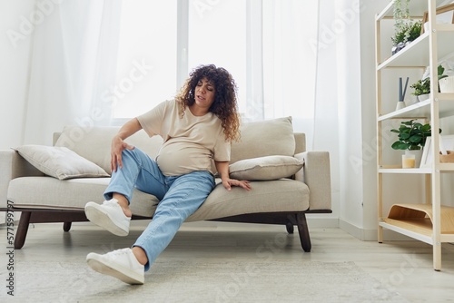 Pregnant woman sitting on sofa at home and putting on pregnancy-friendly shoes with problem due to belly, headache, difficulties of pregnancy and motherhood