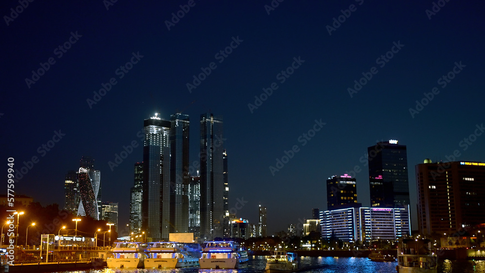 View from the river on the night city with skyscrapers and many lights. Action. Ships and yachts flowing along the river at night.