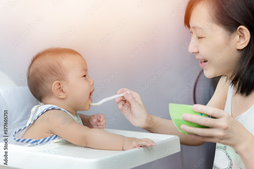Mother feeding baby with spoon, baby boy child sitting eating food on chair at home Mother feeding baby with spoon, baby boy child sitting eating food on chair at home