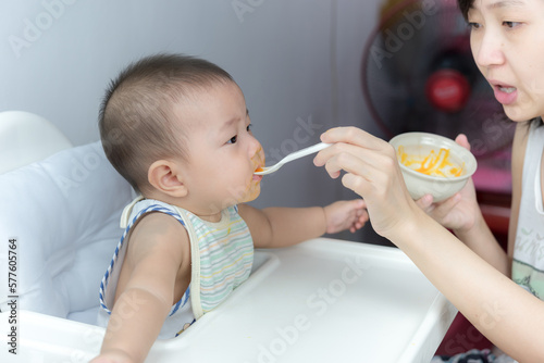 Mother feeding baby with spoon,  baby boy child sitting eating food on chair at home