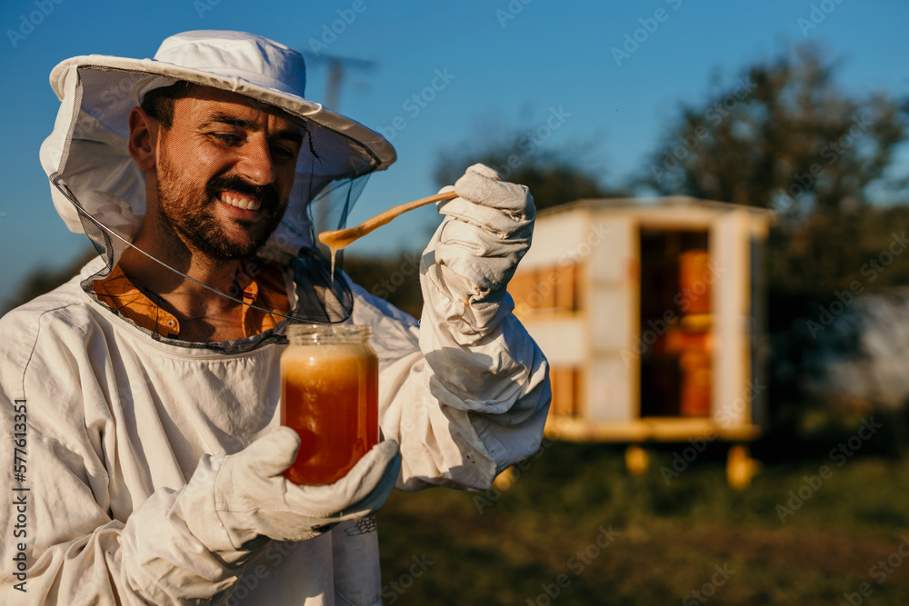A beekeeper worker in his 30s is holding a glass of high-quality bee honey in her hands, wearing gloves and a protective suit, smiling facial expression, focusing on the jar and hand. A beekeeper worker in his 30s is holding a glass of high-quality bee honey in her hands, wearing gloves and a protective suit, smiling facial expression, focusing on the jar and hand.