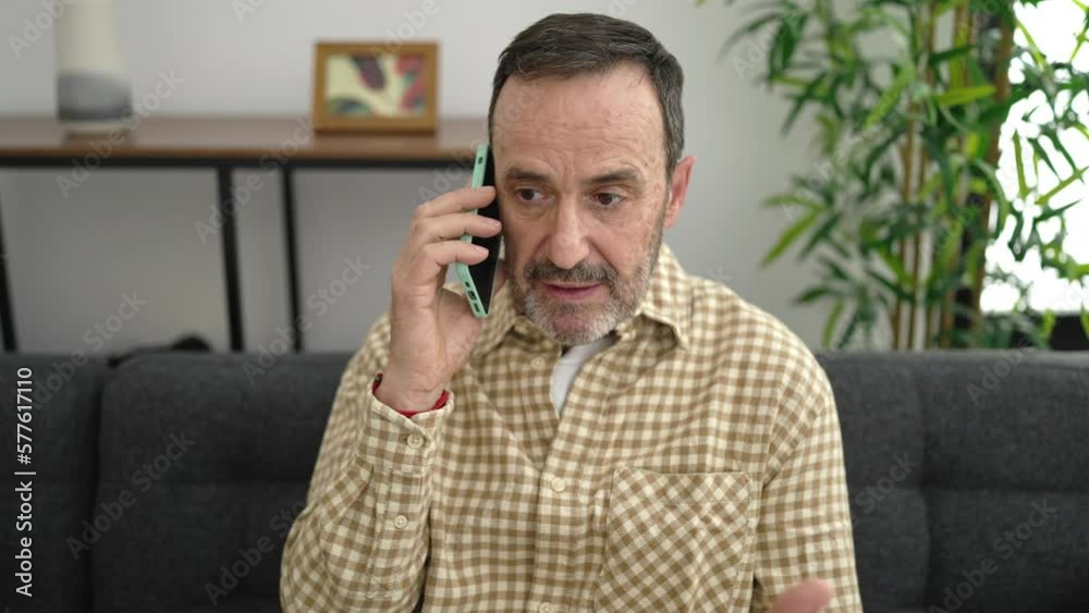 Middle age man arguing by smartphone sitting on sofa at home Middle age man arguing by smartphone sitting on sofa at home