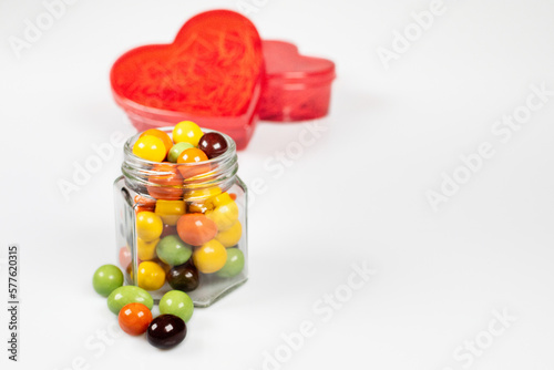 A red heart-shaped translucent gift box and a glass jar with colorful chocolate balls. Confess your love with all your heart