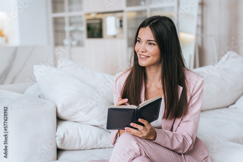 Purposeful brunette Asian young businesswoman in light pink suit holds diary sits on cozy couch looks away dreamily toothy smiles. Cheerful American student girl planning week. Success.