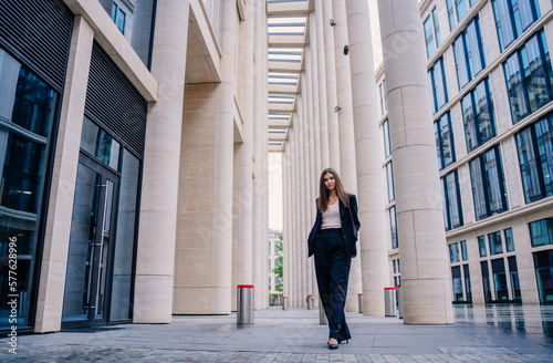 Young brunette businesswoman standing outside looks cat camera. Pretty Asian woman in black suit goes to office. Successful manager against business center. Financial people. Lawyer goes to court.