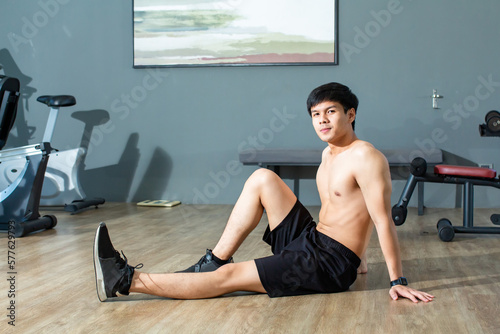 Asian handsome young man is warming up his body by stretching his legs while looking the camera in fitness center.