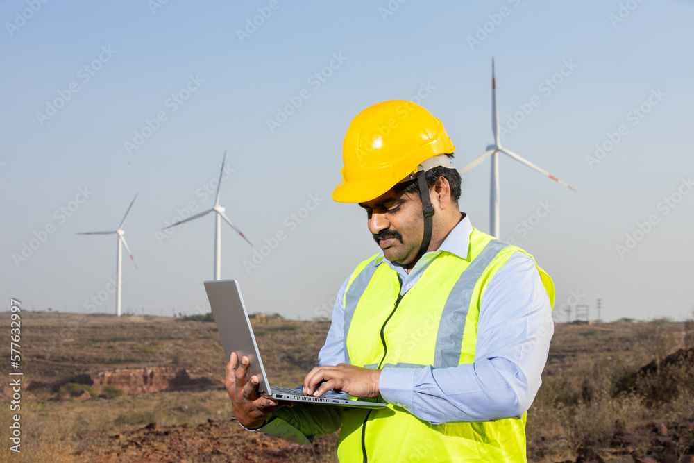 India engineer using laptop to control and maintain windmill farm operation to generate electricity, Asian man wearing yellow helmet and vest working at wind turbine farm, Clean and green energy. India engineer using laptop to control and maintain windmill farm operation to generate electricity, Asian man wearing yellow helmet and vest working at wind turbine farm, Clean and green energy.