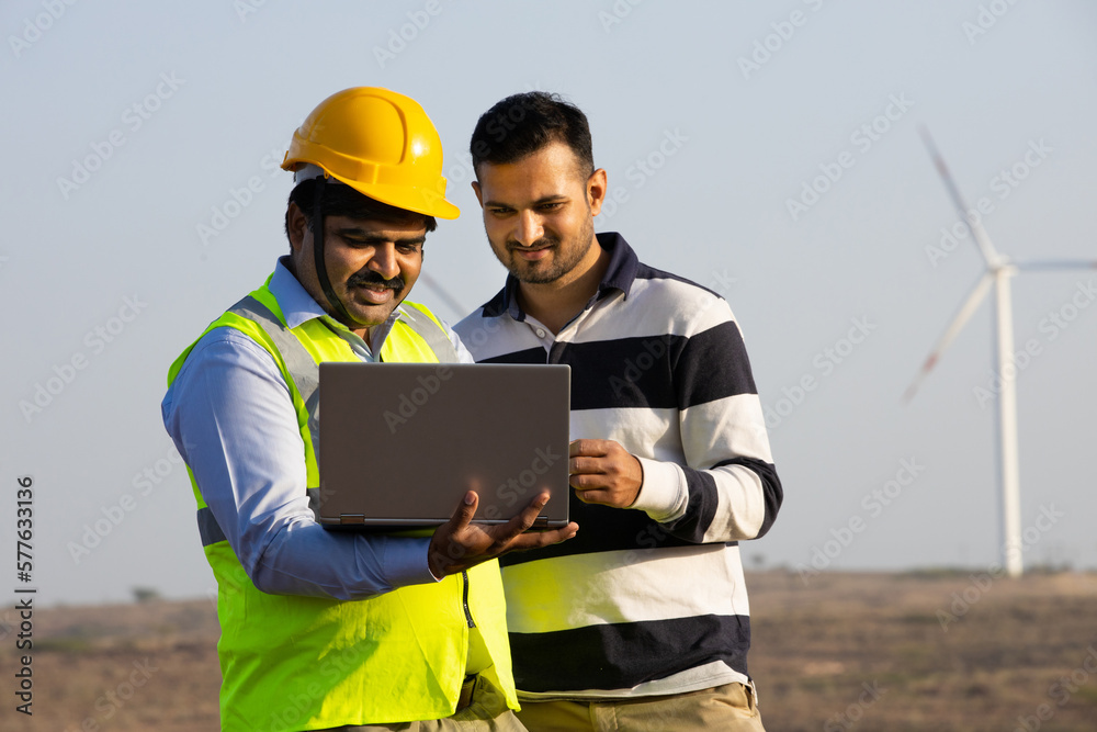 India engineer using laptop and discussing about windmill farm operation to generate electricity, Asian man wearing yellow helmet and vest working at wind turbine farm, clean and green energy. India engineer using laptop and discussing about windmill farm operation to generate electricity, Asian man wearing yellow helmet and vest working at wind turbine farm, clean and green energy.