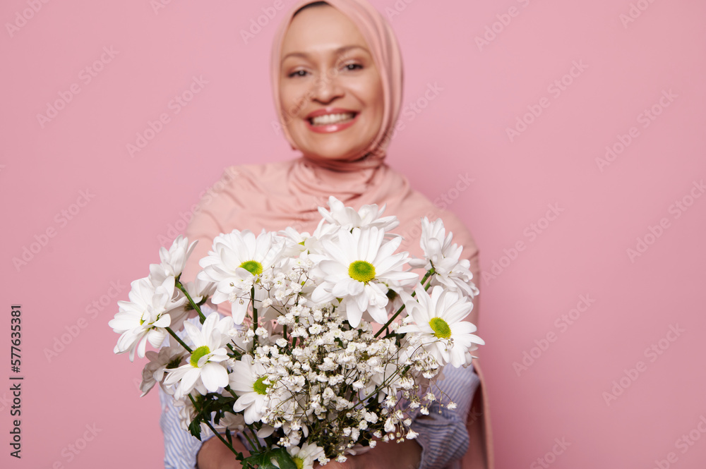Cute spring bouquet of white chamomile or chrysanthemum flowers, in the hands of a blurred Arab Muslim woman, smiling a toothy smile looking at camera, isolated over pink background with copy space Cute spring bouquet of white chamomile or chrysanthemum flowers, in the hands of a blurred Arab Muslim woman, smiling a toothy smile looking at camera, isolated over pink background with copy space