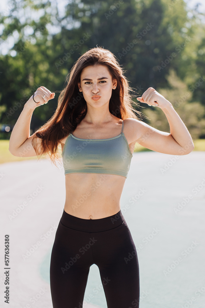 Strong independent young brunette fit girl in sportswear shows double biceps bending hands looks at camera. Fitness, healthy lifestyle. Brunette student at workout outdoors. Sport.