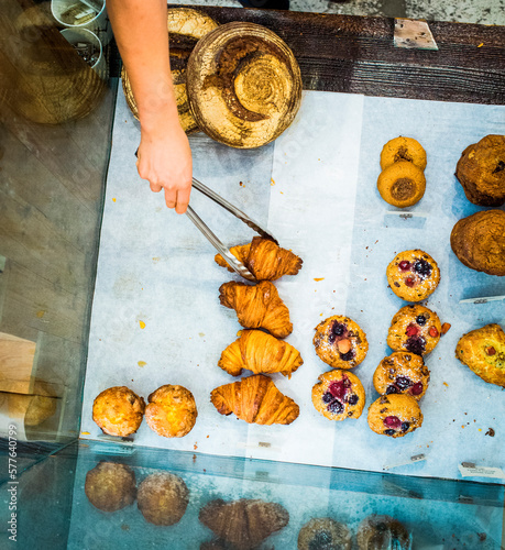 VANCOUVER, BRITISH COLUMBIA, CANADA. A hand reaches into a bakery display case to grab a croissant with silver tongs.