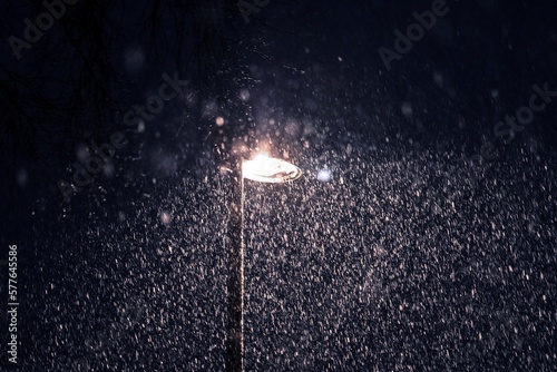 A portrait of a turned on street light or lantern, lighting up a street during a snow storm at night. All the snow flakes are visible in the beam of light during the blue darkness.