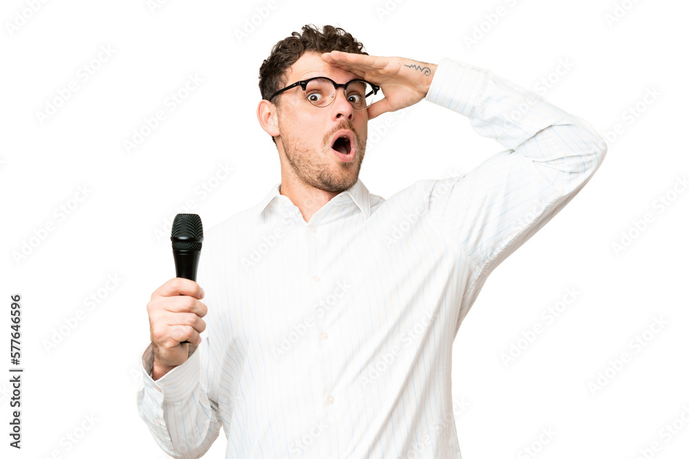 Brazilian man picking up a microphone over isolated chroma key background doing surprise gesture while looking to the side Brazilian man picking up a microphone over isolated chroma key background doing surprise gesture while looking to the side
