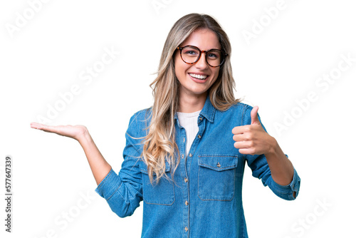 Young Uruguayan woman over isolated background holding copyspace imaginary on the palm to insert an ad and with thumbs up
