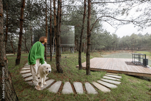 Woman walks on wavy pathway near lake and invisible cabin in pine forest. Connection with nature and sustainability concept. Rest in tiny cabins on nature