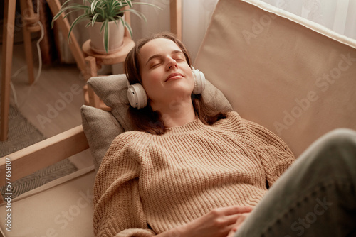 Happy Caucasian woman lying on sofa and listening to music in headphones, enjoying audio playing in earphones, relaxing and chilling at home, expressing positive emotions having fun while laying along