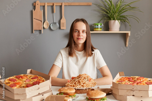 Indoor shot of calm attractive woman with brown hair wearing white T-shirt sitting at table among different fast food  looking at camera  being ready to have dinner with junk dish.