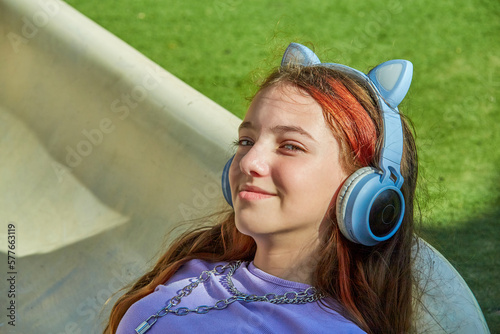 A cute, beautiful teenage girl in blue headphones with cat ears lies across a children's slide in a children's playground and listens to music on a sunny warm day