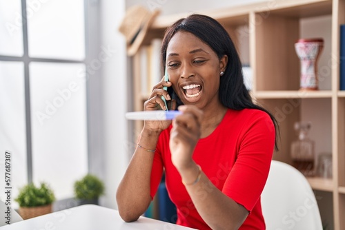 African young woman holding pregnancy test result speaking on the phone smiling and laughing hard out loud because funny crazy joke.