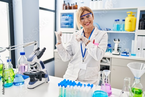 Middle age blonde woman working at scientist laboratory pointing to the back behind with hand and thumbs up  smiling confident