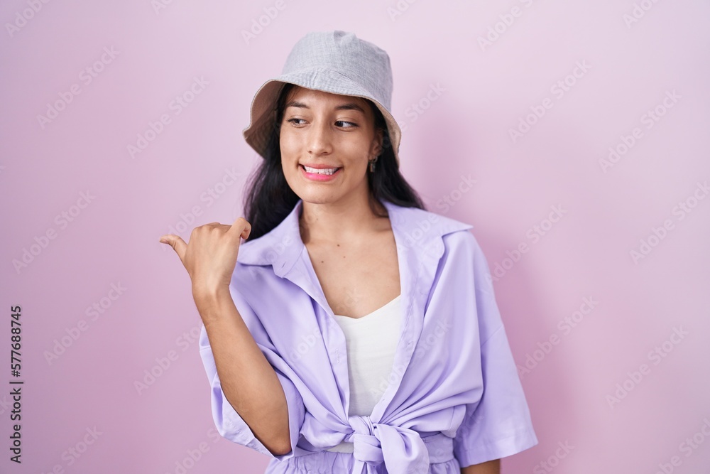Young hispanic woman standing over pink background wearing hat smiling with happy face looking and pointing to the side with thumb up. Young hispanic woman standing over pink background wearing hat smiling with happy face looking and pointing to the side with thumb up.