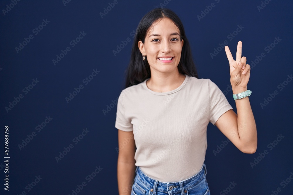 Young hispanic woman standing over blue background showing and pointing up with fingers number two while smiling confident and happy. Young hispanic woman standing over blue background showing and pointing up with fingers number two while smiling confident and happy.