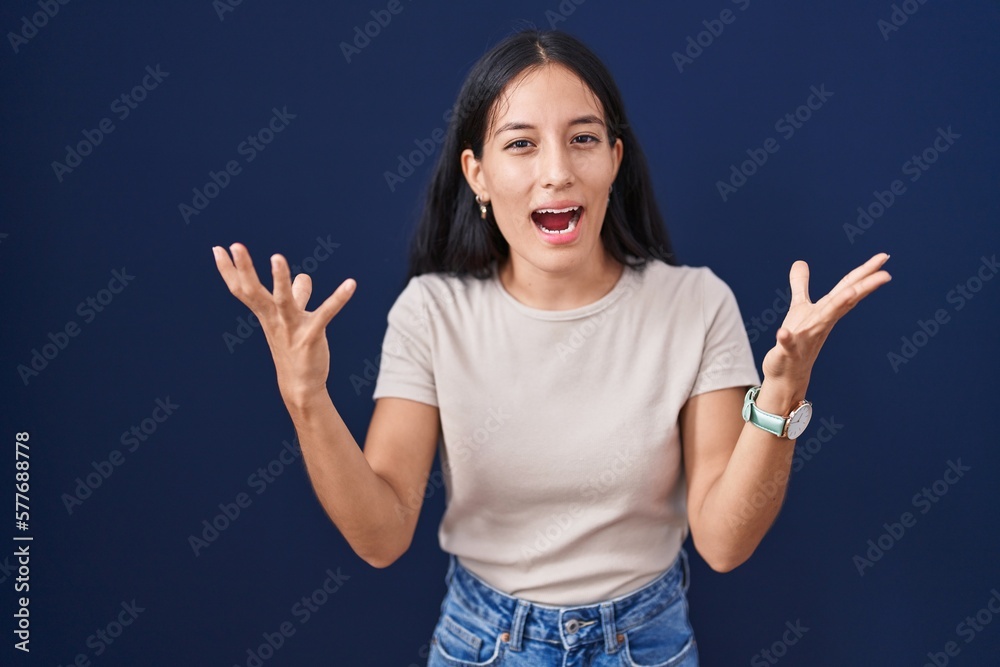 Young hispanic woman standing over blue background crazy and mad shouting and yelling with aggressive expression and arms raised. frustration concept.