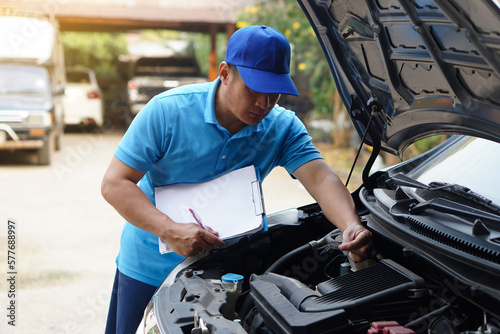 Asian man mechanic wears blue cap and blue shirt, holds paper notepad, checking and analyzing car engine under the hood, found insect that causes engine broken .Concept, Outdoor car inspection service