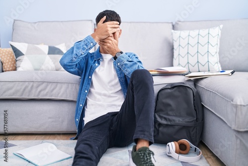 Young hispanic man sitting on the floor studying for university covering eyes and mouth with hands, surprised and shocked. hiding emotion