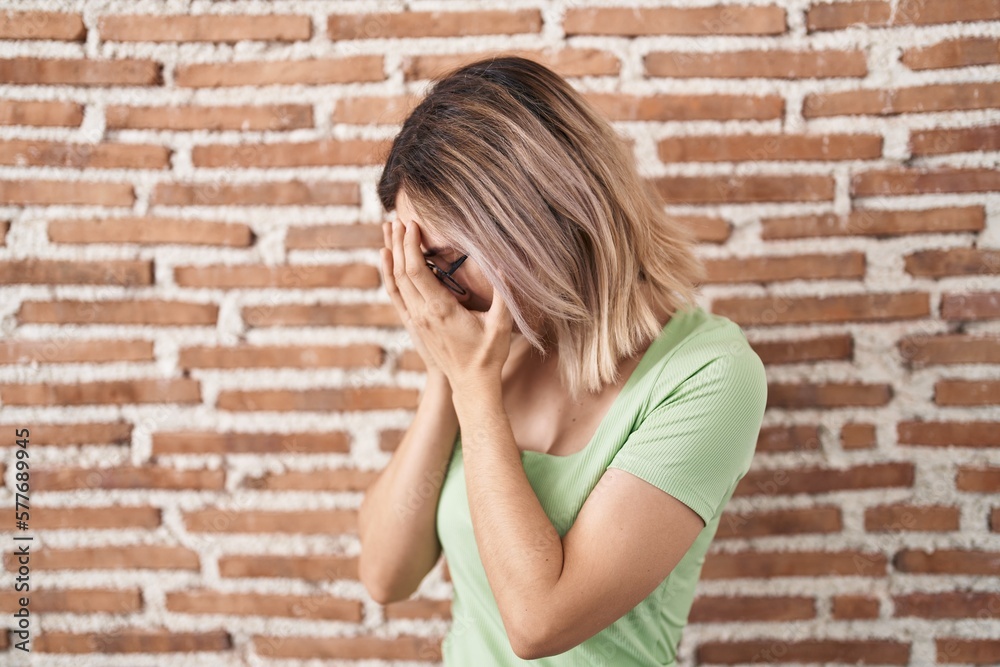 Young beautiful woman standing over bricks wall with sad expression covering face with hands while crying. depression concept.