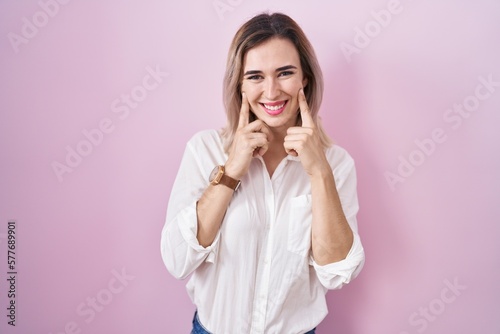 Young beautiful woman standing over pink background smiling with open mouth, fingers pointing and forcing cheerful smile