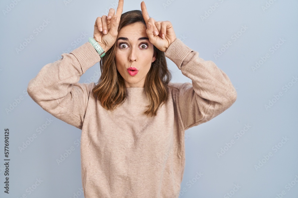 Fototapeta premium Young woman standing over isolated background doing funny gesture with finger over head as bull horns
